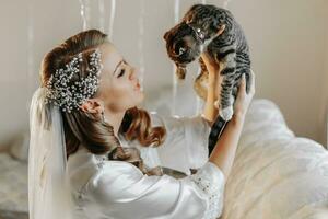 jeune, élégant, fille dans le image de une la mariée avec une magnifique agrafe et une tiare sur sa diriger, en portant sa animal de compagnie chat dans sa bras photo