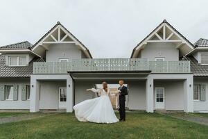 une couple dans l'amour sur le Contexte de gris Maisons. une blond la mariée dans une mariage robe avec manches jette en haut sa robe. jeune marié dans une classique noir costume, blanc chemise et cravate. sourit et joie photo