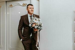 le jeune marié des stands dans le pièce avec une bouquet de fleurs dans une classique marron costume et blanc chemise. préparation pour le mariage la cérémonie photo