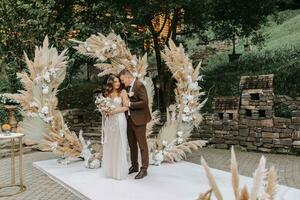 le la mariée et jeune marié sont profiter eux-mêmes. jeunes mariés avec une mariage bouquet permanent à une mariage la cérémonie en dessous de un cambre décoré avec fleurs et séché fleurs en plein air. photo