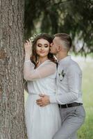 portrait de le la mariée et jeune marié. une la mariée avec longue cheveux et une couronne sur sa tête près une grand arbre, de derrière le jeune marié câlins sa et baisers sa sur le joue. photo