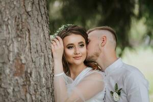 portrait de le la mariée et jeune marié. une la mariée avec longue cheveux et une couronne sur sa tête près une grand arbre, de derrière le jeune marié câlins sa et baisers sa sur le joue. photo