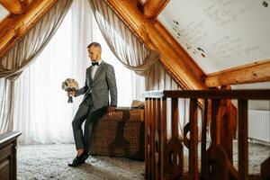 une Jeune jeune marié dans une Hôtel pièce portant une classique gris costume, blanc chemise et arc cravate. du repos dans magnifique appartements. le du marié Matin à le Hôtel. Masculin mains proche en haut photo