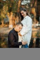 Jeune Enceinte femme dans une blanc chemise. le mari mettre le sien mains sur le sien femme estomac et penché contre le sien oreille. photo