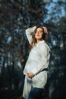 une Enceinte femme dans une blanc robe sur une foncé Contexte pose dans le parc. content et insouciant grossesse. l'automne parc. content femme profiter sa grossesse. photo