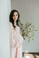 une magnifique Enceinte femme dans une rose robe détient sa mains sur sa estomac. une aimant mère est attendre pour le naissance de une enfant. concept de grossesse, maternité, préparation et en attendant. photo