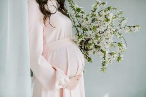 fermer photo de une Enceinte femme dans une rose robe en portant sa mains sur sa estomac. une aimant mère est attendre pour le naissance de une enfant. le concept de grossesse, maternité.