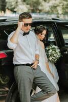 mariage portrait, photo de une élégant jeune marié dans une blanc chemise et arc attacher et une brunette la mariée avec une bouquet de fleurs près une noir auto.