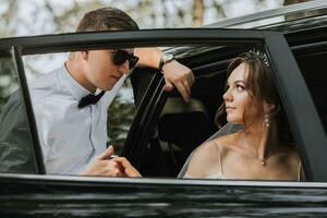 mariage portrait, photo de une élégant jeune marié dans une blanc chemise et arc attacher et une brunette la mariée avec une bouquet de fleurs près une noir auto.