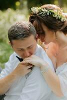une magnifique mignonne la mariée dans une robe câlins derrière le retour de une élégant jeune marié dans une blanc chemise qui est séance sur vert herbe. mariage portrait, photo de jeunes mariés dans l'amour.