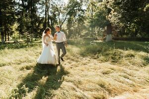 une Jeune couple de jeunes mariés sont en marchant dans une été parc. le la mariée dans une magnifique blanc robe avec une couronne de Frais fleurs sur sa diriger, le jeune marié dans une blanc chemise photo