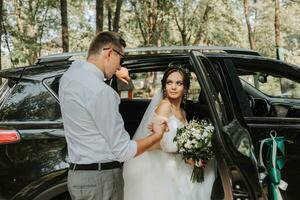 mariage portrait, photo de une élégant jeune marié dans une blanc chemise et arc attacher et une brunette la mariée avec une bouquet de fleurs près une noir auto.