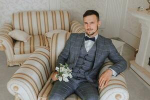 une élégant barbu jeune marié avec une bouquet de fleurs est séance sur une chaise dans un coûteux Hôtel chambre. du marié Matin. le jeune marié est avoir prêt dans le Matin avant le mariage la cérémonie photo