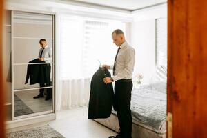 attrayant Jeune homme pose comme une jeune marié avant le mariage, en mettant sur une veste. le jeune marié est en train de préparer pour le mariage dans le chambre photo