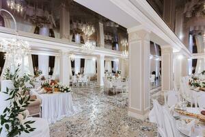 le intérieur de le mariage salle dans blanc couleurs, le les tables sont ensemble, tout est décoré avec fleurs, cristal lustres accrocher, le réflexion sur le plafond ajoute le volume photo