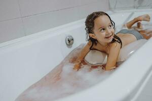 petit, souriant, magnifique aux cheveux noirs fille avec longue cheveux, enfant baignades, lavages dans une blanc une baignoire avec mousse. amusement photo. photo