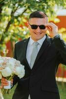 photo de le jeune marié dans une classique noir costume et lunettes, avec une bouquet de fleurs dans le sien mains