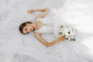 magnifique la mariée avec mariage bouquet de fleurs, attrayant femme dans mariage robe. le la mariée rêveusement mensonges sur sa robe, le photo est pris de au dessus