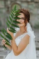 mariage portrait. le la mariée dans une blanc robe est poser, en portant une feuille près sa visage et à la recherche dans le lentille. agréable se maquiller. portrait de le la mariée photo