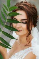 mariage portrait. le la mariée dans une blanc robe est poser, en portant une feuille près sa visage et à la recherche dans le lentille. agréable se maquiller. portrait de le la mariée photo