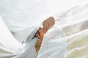 mariage portrait. le la mariée et jeune marié couvert se avec une voile, étreint et embrassé tendrement. vacances concept. transparent voile. des rayons de le Soleil. photo