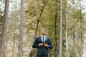 une Jeune homme dans une classique costume contre le Contexte de grand des arbres dans une forêt ou parc. le jeune marié attache le sien veste photo