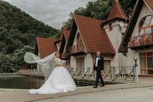 une brunette la mariée dans une longue robe et une jeune marié dans une classique costume sont permanent sur une pont près une Lac contre le Contexte de une château. une voile est jeté dans le air photo