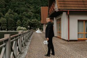 mariage portrait. une homme dans une noir costume des stands sur une pont avec une bouquet et regards loin. fiancé. masculin, affaires style photo