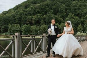 une brunette la mariée dans une longue robe et une jeune marié dans une classique costume sont en marchant sur une pont près une Lac contre le Contexte de une château. une marcher dans la nature. mariage journée photo