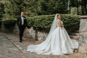 mariage portrait. le la mariée dans un élégant robe des stands dans de face de le jeune marié dans une classique costume, contre le Contexte de vert des arbres. longue train. été mariage. une marcher dans la nature photo