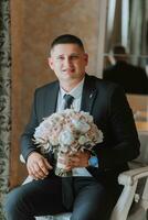 une élégant jeune marié avec une bouquet de fleurs est séance sur une chaise dans un coûteux Hôtel chambre. du marié Matin. le jeune marié est avoir prêt dans le Matin avant le mariage la cérémonie photo