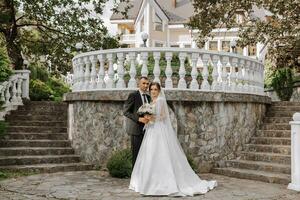 portrait de une Jeune la mariée dans une blanc robe et une brunette jeune marié dans une costume sur le escaliers embrassement près un vieux ville parc. une magnifique et romantique mariage, une content couple photo