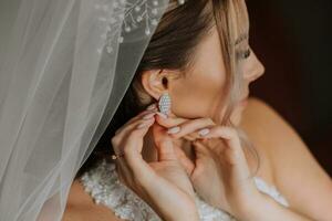 une magnifique la mariée attache un boucle d'oreille. fermer portrait de une magnifique fille dans une mariage robe dans le intérieur. mariage la photographie photo