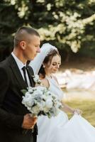 une élégant jeune marié dans une noir costume et une mignonne la mariée dans une blanc robe avec une longue voile sont étreindre dans une parc. mariage portrait de souriant et content jeunes mariés. photo