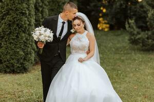 une élégant jeune marié dans une noir costume et une mignonne la mariée dans une blanc robe avec une longue voile sont étreindre dans une parc. mariage portrait de souriant et content jeunes mariés. photo