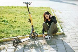 Extérieur portrait de Jeune brunette adolescent fille avec court cheveux attacher les lacets près scooter dans ville carré photo