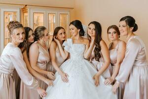 élégant magnifique demoiselles d'honneur dans correspondant à soie Robes portion magnifique brunette la mariée dans blanc robe avoir prêt pour mariage, Matin les préparatifs, femme en mettant sur robe photo