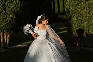 soumissionner portrait de une content la mariée avec une bouquet de orchidées dans sa mains, en plein air. chaud été temps. une longue voile, une luxueux blanc robe avec une train. photo