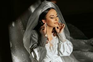 magnifique la mariée avec longue foncé frisé cheveux dans une longue blanc peignoir dans le Matin. le la mariée est en train de préparer pour le mariage. professionnel maquillage et cheveux. une couronne sur le tête et une longue voile de le la mariée photo