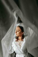 magnifique la mariée avec longue foncé frisé cheveux dans une longue blanc peignoir dans le Matin. le la mariée est en train de préparer pour le mariage. professionnel maquillage et cheveux. une couronne sur le tête et une longue voile de le la mariée photo