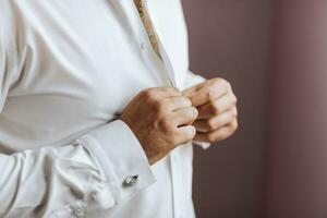 fermer de une homme boutonnage le sien chemise. le jeune marié est en train de préparer pour le mariage. le homme porte une blanc chemise. élégant jeune marié photo