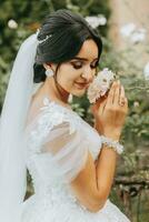 portrait de le la mariée dans une blanc robe avec une voile sur sa diriger. professionnel mariage maquillage et cheveux. magnifique Jeune la mariée. une content femme jouit le arôme de fleurs photo