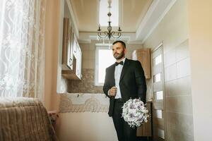 une élégant jeune marié est en train de préparer pour le mariage la cérémonie dans le Matin. du marié Matin. une homme dans une classique costume et une arc cravate. Jeune et Beau jeune marié avec une bouquet de fleurs photo