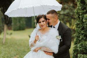 la mariée et jeune marié avec une blanc parapluie, des promenades dans le pluie dans le parc, mariage photo session dans le pluie. une modèle avec un parapluie dans une magnifique mariage robe. le jeune marié pistes le la mariée par le main