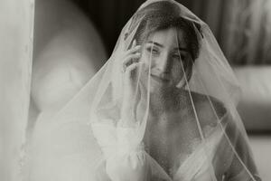 une étourdissant Jeune la mariée pose par le fenêtre en portant sa voile dans sa mains. une magnifique femme de caucasien apparence dans un élégant blanc peignoir et luxueux coiffure. mariage maquillage photo