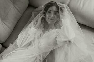 une magnifique la mariée est mensonge sur une moderne canapé dans une blanc peignoir. mariage coiffure, professionnel se maquiller. romantique la mariée posant photo