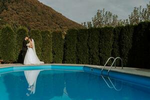 portrait de une la mariée et jeune marié embrasser dans un infini bassin contre le ciel et haute montagnes photo