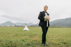 une Beau jeune marié dans une noir costume et une noir arc attacher des stands en plein air dans une large champ contre une Contexte de montagnes. mariage portrait. une homme dans une classique costume dans le premier plan. photo