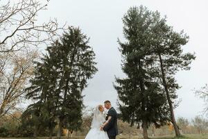 content élégant couple de jeunes mariés dans le vert forêt sur un l'automne journée. le la mariée dans une classique longue blanc robe et le jeune marié dans une bleu costume embrasser. mariage journée. photo