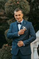 une Beau jeune marié dans une bleu costume et noir attacher est permanent en plein air. mariage portrait. une homme dans une classique costume contre le Contexte de l'automne feuilles photo